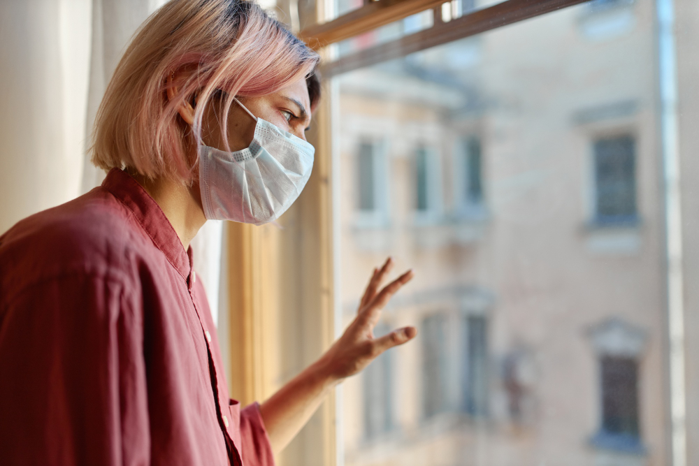 teenage girl with pinkish hair standing in front of closed window with hand on glass looking outside while staying at home during quarantine coronavirus pandemic and social distancing concept